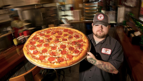 A chef holds a large pepperoni pizza in a busy restaurant kitchen.