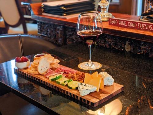 A wooden charcuterie board with meats, cheeses, bread, cucumbers, grapes, and a glass of red wine on a dark table.