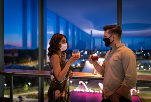 A couple in masks clinks glasses while enjoying drinks at a stylish venue with a city view at dusk.