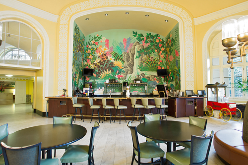A vibrant bar area with tropical mural, surrounded by round tables and chairs in a bright, airy space.