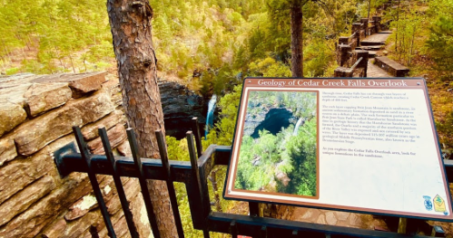 Sign about the geology of Cedar Creek Falls Overlook, with a view of the falls and surrounding trees.
