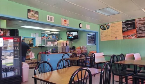 Interior of a casual eatery with tables, a counter, and a TV, featuring a menu on the wall and a fridge in the corner.