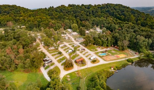 Aerial view of a campground with RVs, a pool, and a pond, surrounded by lush green trees and hills.