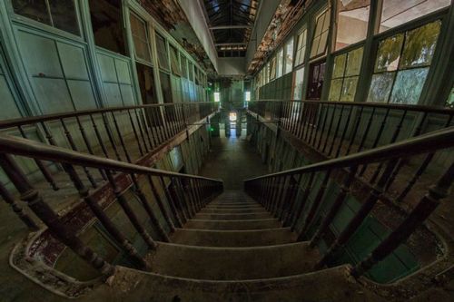 A dimly lit, abandoned staircase leads down through an old building with peeling walls and green-tinted light.