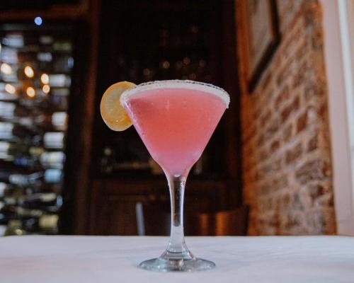 A pink cocktail in a martini glass, garnished with a lemon slice and rimmed with salt, set against a rustic background.