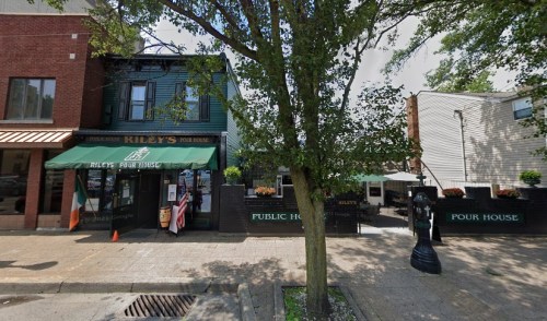 Exterior view of Riley's Pour House, a pub with outdoor seating, surrounded by trees and nearby buildings.
