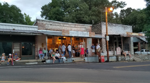 A group of people gathers outside a rustic storefront in a small town, with trees and vintage buildings in the background.