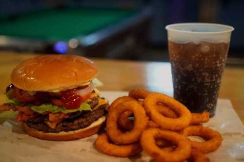 A juicy burger with lettuce, tomato, and bacon, served with onion rings and a cup of soda on a wooden table.