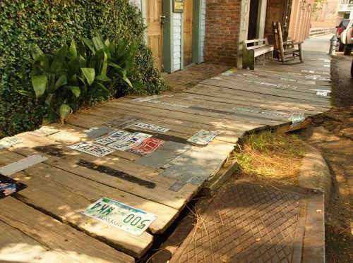 A wooden walkway with various license plates embedded, surrounded by greenery and brick buildings.