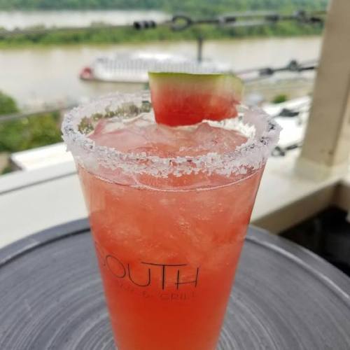 A refreshing pink cocktail with a salted rim and a watermelon slice, served on a tray with a scenic river view.