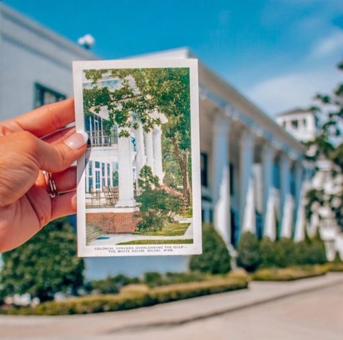 A hand holds a vintage postcard of a colonial building with columns, set against a bright blue sky.