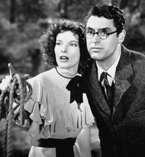 A black-and-white scene featuring a woman and a man looking intently off-screen, surrounded by trees.