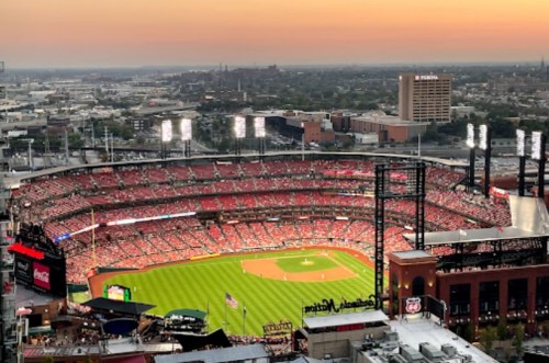 Aerial view of a packed baseball stadium at sunset, with city skyline in the background.