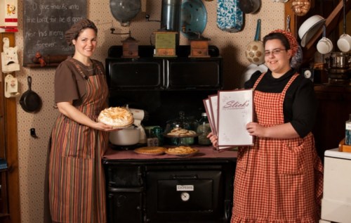 Two women in vintage aprons stand in a rustic kitchen, one holding a dessert and the other a menu, with baked goods nearby.