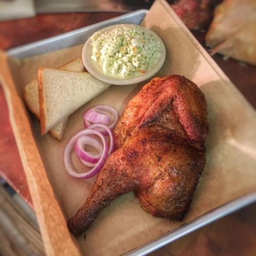 A plate featuring smoked chicken leg, coleslaw, two slices of bread, and onion rings on parchment paper.