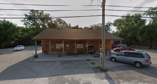 A wooden building with a sloped roof, surrounded by trees and parked cars, featuring outdoor seating.