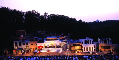 A vibrant outdoor theater stage at dusk, with a large audience and scenic backdrop of buildings and trees.