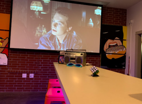 A retro scene with a movie playing on a wall, a boombox, sunglasses, and a Rubik's Cube on a bar counter.