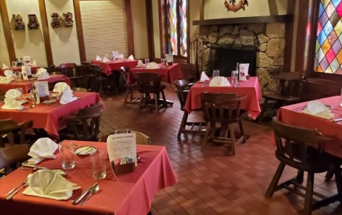 Cozy restaurant interior with red tablecloths, wooden chairs, and a stone fireplace, featuring stained glass windows.