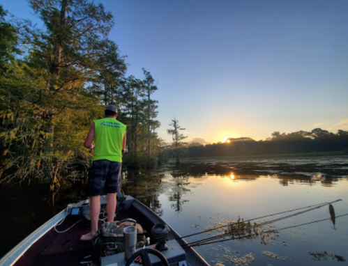 A person fishing from a boat at sunset, surrounded by trees and calm water.
