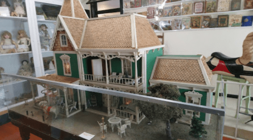 A detailed dollhouse with a green exterior, multiple stories, and a porch, displayed in a museum setting.