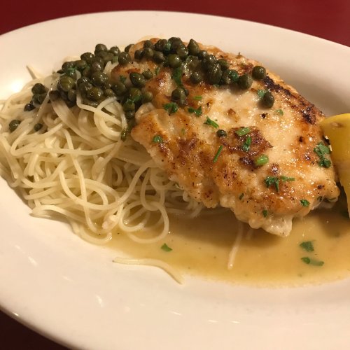 Grilled fish fillet topped with capers, served over a bed of pasta with a lemon wedge on the side.