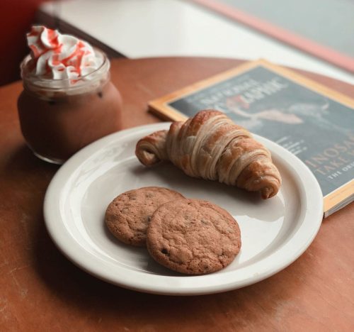 A cozy café scene featuring a glass of iced chocolate, a croissant, and two cookies on a plate beside a magazine.