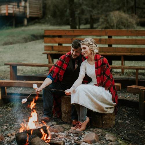 A couple in plaid blankets sits by a campfire, roasting marshmallows in a cozy outdoor setting.