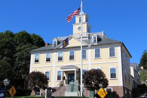 Historic yellow building with a clock tower, American flag, and a statue in front, surrounded by trees and a clear blue sky.