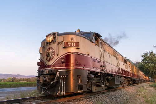 A vintage train with a maroon and silver exterior travels along a scenic route, emitting steam against a clear sky.
