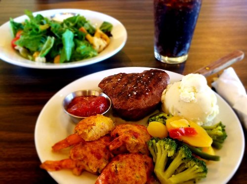 A plate with steak, shrimp, mashed potatoes, and mixed vegetables, alongside a salad and a glass of cola.