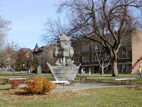 A large stone sculpture of a figure holding an object, surrounded by trees and a grassy area on a campus.