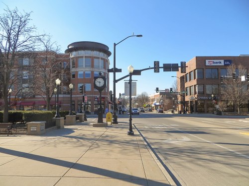 A city street view featuring a clock tower, bank, and shops under a clear blue sky. Traffic lights and trees are visible.