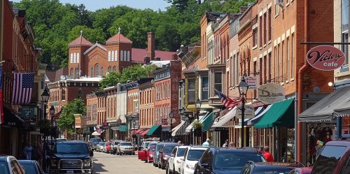 A charming street lined with brick buildings, shops, and parked cars, set against a backdrop of green hills.