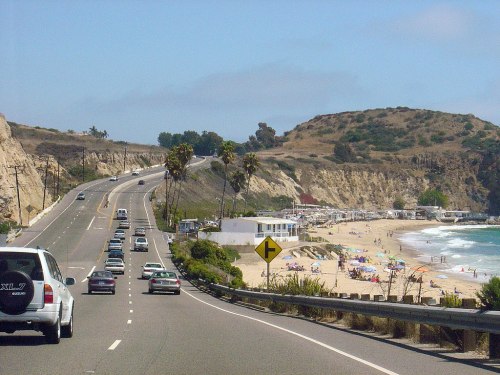 A scenic coastal road with cars, palm trees, and a beach in the background under a clear blue sky.