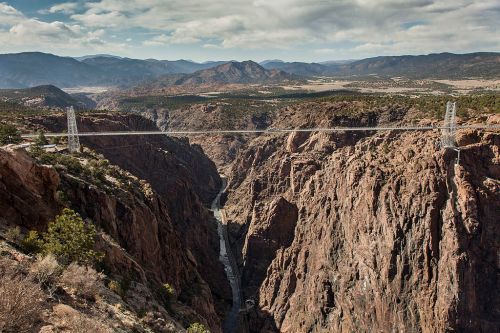A suspension bridge spans a deep canyon, surrounded by rugged mountains and a cloudy sky.