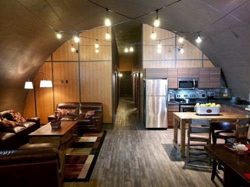 Modern, spacious living area with leather sofas, wooden accents, and a kitchen, illuminated by string lights.