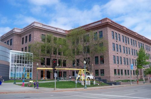 Historic brick building with large windows, surrounded by trees, and a modern glass structure nearby. Clear blue sky above.