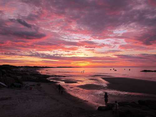 A vibrant sunset over a beach, with colorful clouds reflecting on the water and silhouettes of people walking along the shore.