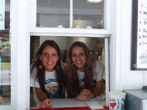 Two young women smile from a service window, wearing matching t-shirts and earrings, with a menu visible behind them.