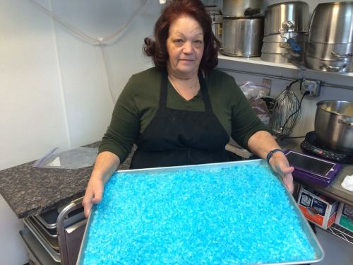 A woman in an apron holds a tray filled with bright blue crystals in a kitchen setting.