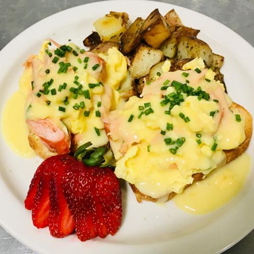 A plate of eggs Benedict with salmon, hollandaise sauce, chives, roasted potatoes, and a strawberry on the side.