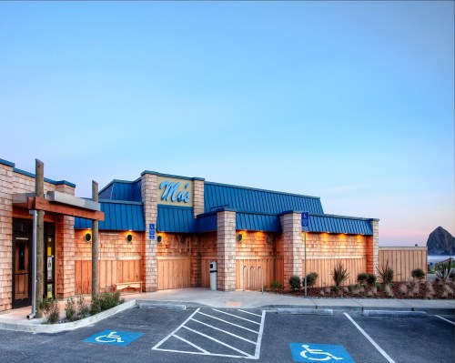 A wooden building with a blue roof, labeled "M's," set against a clear sky and parking spaces, including accessible spots.
