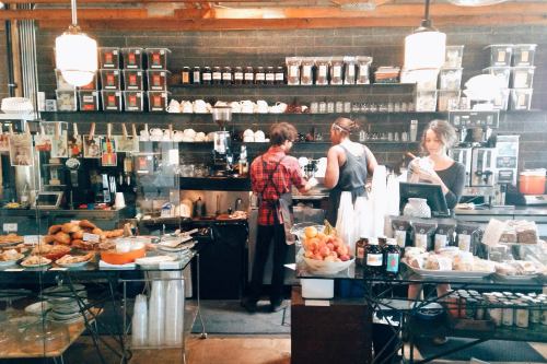 A busy café scene with baristas preparing drinks, pastries on display, and shelves filled with coffee products.