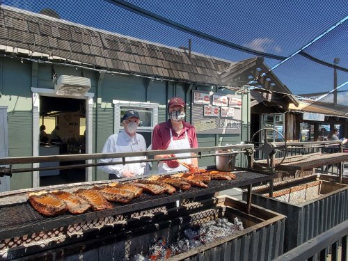 Two chefs in masks stand by a grill, cooking ribs outdoors at a restaurant with a sunny sky above.