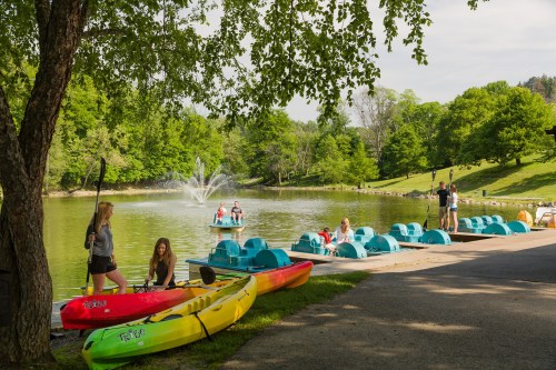 People enjoying a sunny day by a lake, with kayaks and paddle boats, surrounded by lush greenery and a fountain.