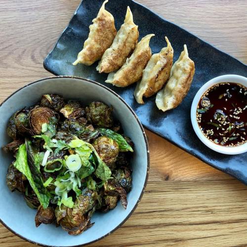 A bowl of crispy Brussels sprouts with green onions, alongside a plate of dumplings and a dipping sauce.