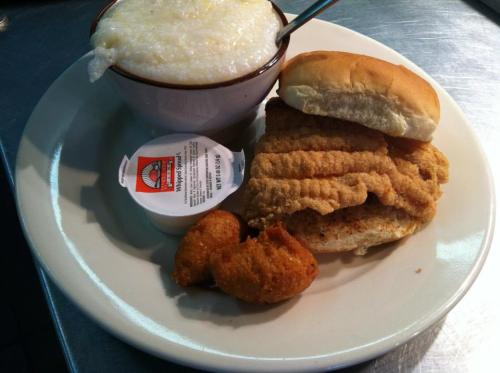 A plate with fried fish sandwich, a bowl of grits, and a side of hushpuppies with a dipping sauce.