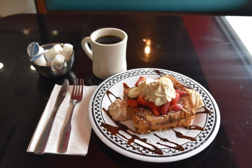 A plate of French toast topped with strawberries, bananas, whipped cream, and chocolate sauce, alongside a cup of coffee.