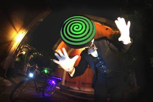 A figure in a suit with a spiral-patterned head and white gloves poses dramatically in a dimly lit outdoor setting.